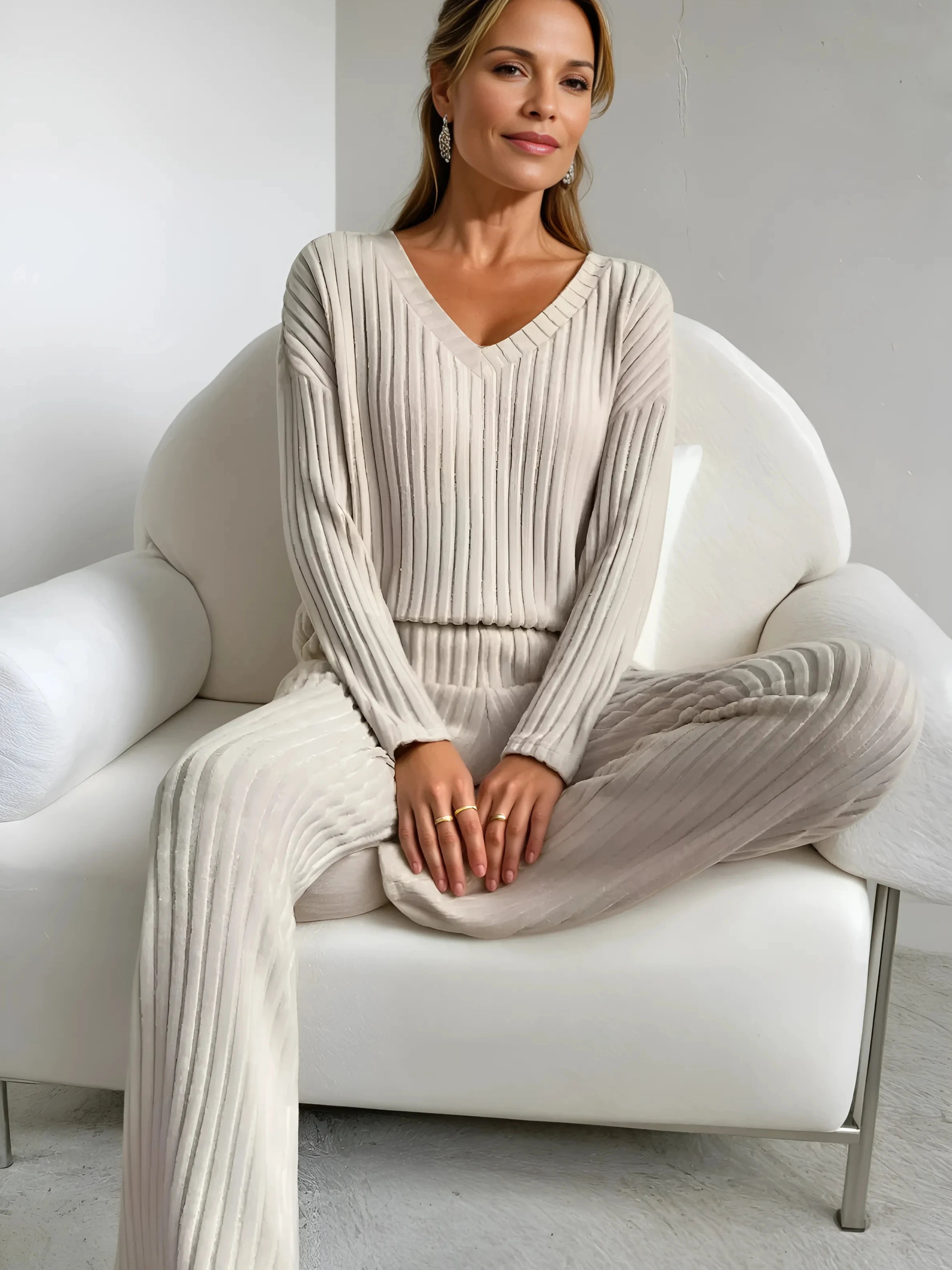 Femme assise sur un fauteuil blanc, vêtue d’un ensemble homewear beige plissé avec un col en V. Une tenue confortable et élégante pour un moment de détente avec style.