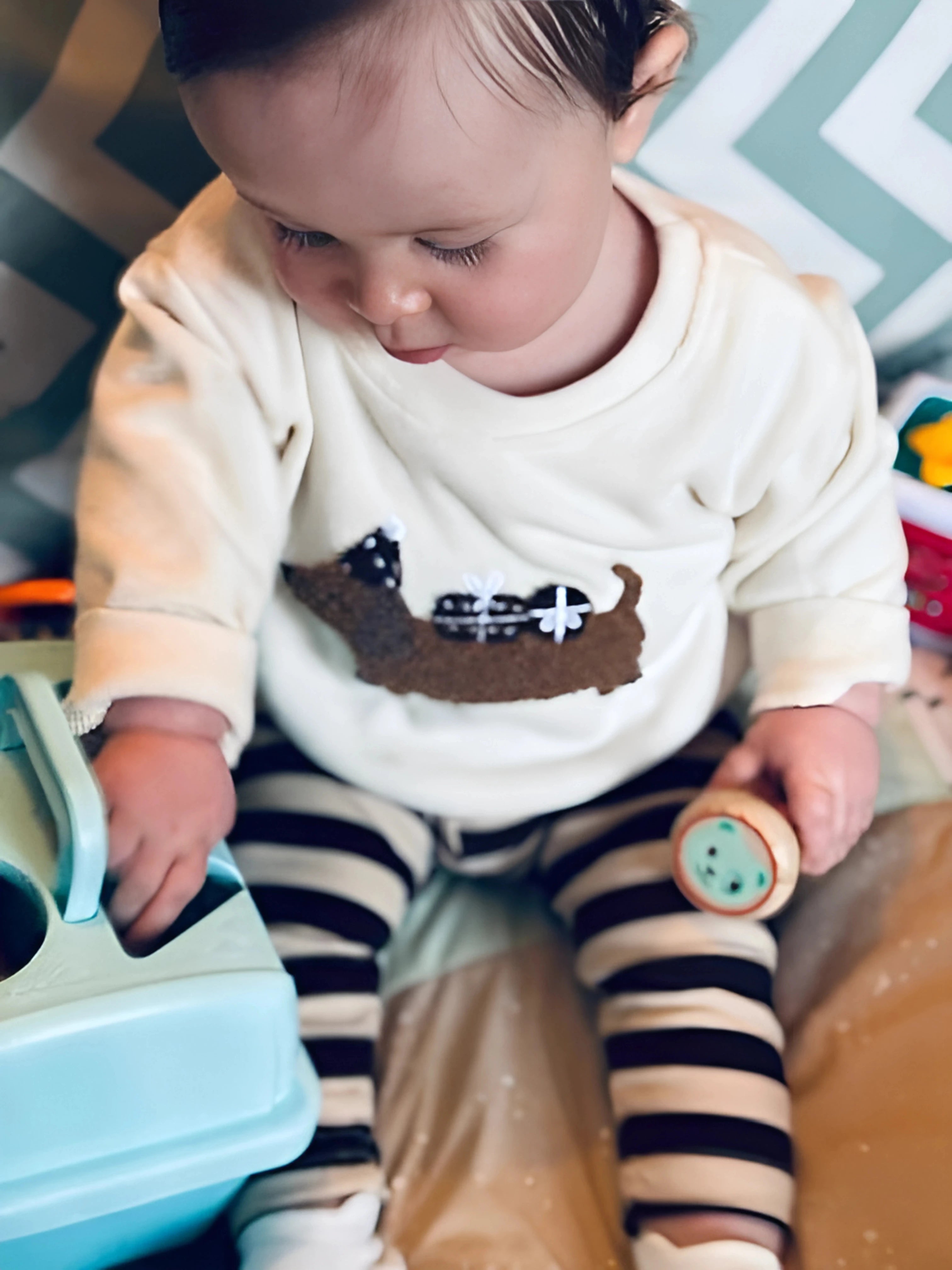 Gros plan sur un bébé curieux jouant avec son jouet, habillé d’un pull en maille crème avec une broderie chien et d’un legging noir et blanc à rayures.